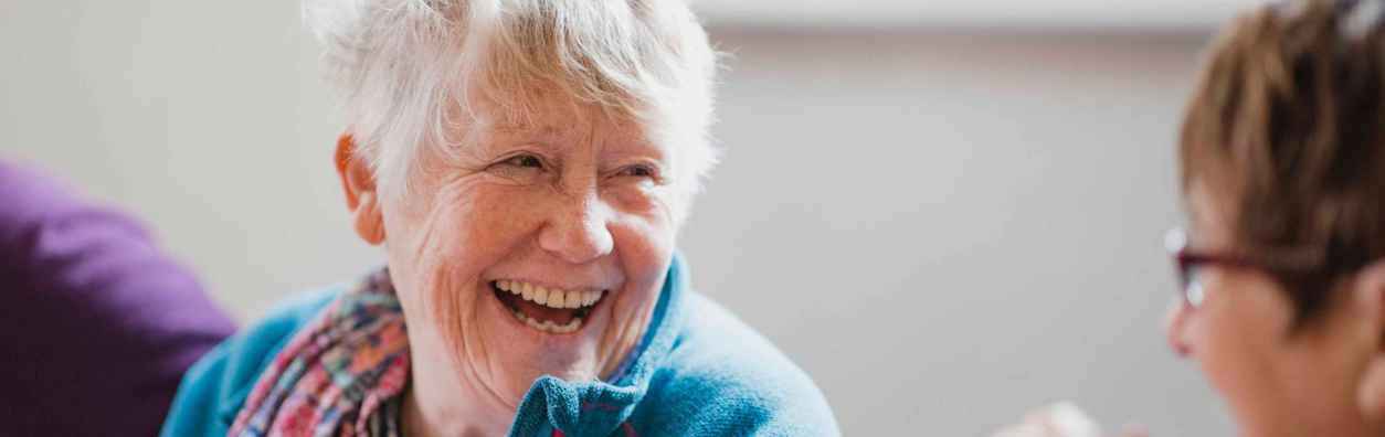 Close-up of an older woman with short grey hair smiling widely at a woman with short dark hair wearing glasses.