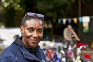 Woman smiling directly at us. Behind her the background is blurred but we can make out a cyclist, trees and colourful flags hanging between the branches.