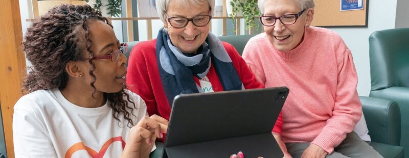 Image of three women looking at a laptop. Far left the person is wearing a Sky Up t shirt and is holding the device and showing the two other women, who are older and sat on a sofa, the screen.