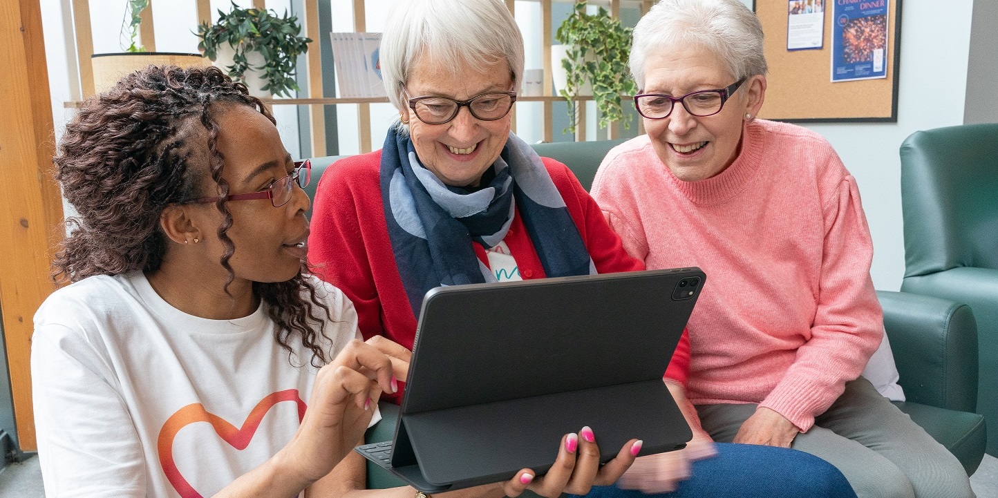 Image of three women looking at a laptop. Far left the person is wearing a Sky Up t shirt and is holding the device and showing the two other women, who are older and sat on a sofa, the screen.