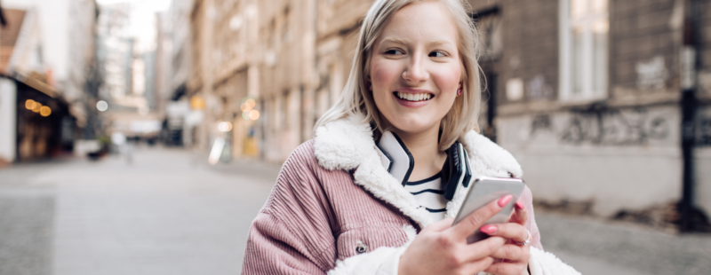 Young woman holding a phone and smiling as she walks outside.