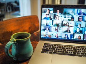 image of coffee cup on table next to laptop, screen filled with faces on an online meeting.