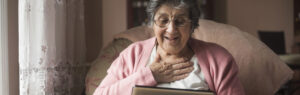 Older woman holding her left hand to her chest and smiling as she looks down at an ipad she is holding with her right hand.