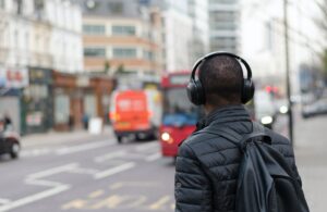 The back of a man wearing headphones and a black puffer jacket and bag. He is waiting to cross a busy road.