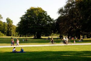A sunny park with people sitting on the grass. There is a path with people cycling and walking on.