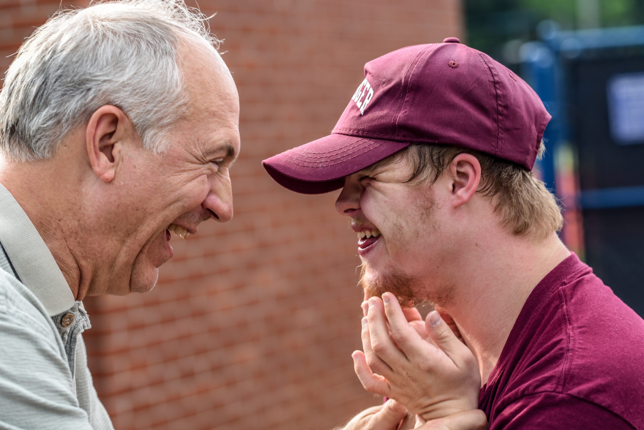 older man with grey hair smiling at younger man with a learning disability in a baseball cap who is also smiling with their hands up to their chin.